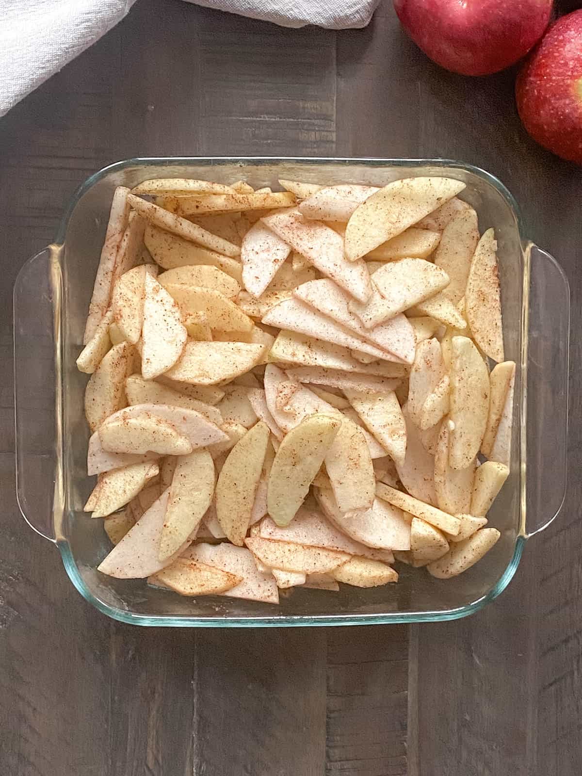 Peeled apple slices in an 8x8 inch glass baking dish covered in cinnamon, maple syrup and water.