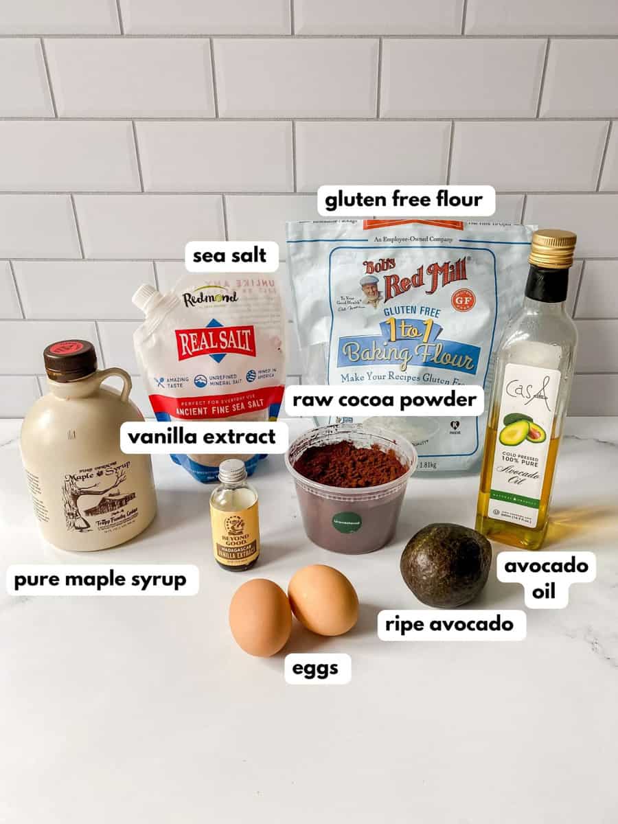 Brownie ingredients set out on counter - pure maple syrup, vanilla extract, sea salt, gluten free flour, raw cocoa powder, 2 eggs, ripe avocado, avocado oil. 
