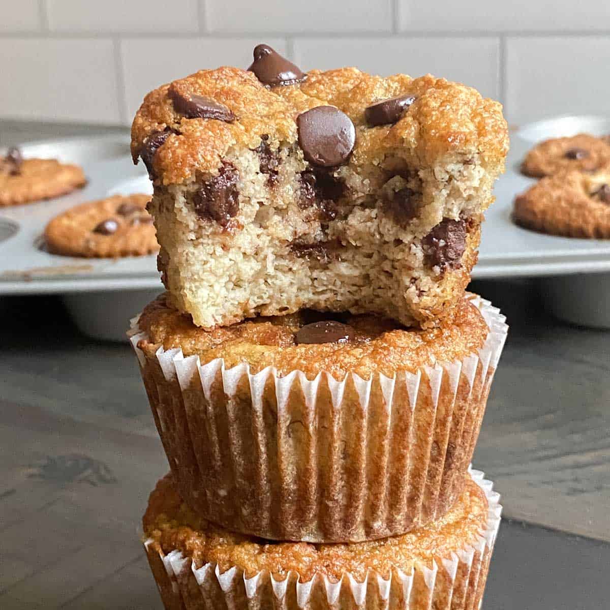 three banana muffins with chocolate chips stacked on top of each other, the top one has a bite taken out of it to show the fluffy inside.