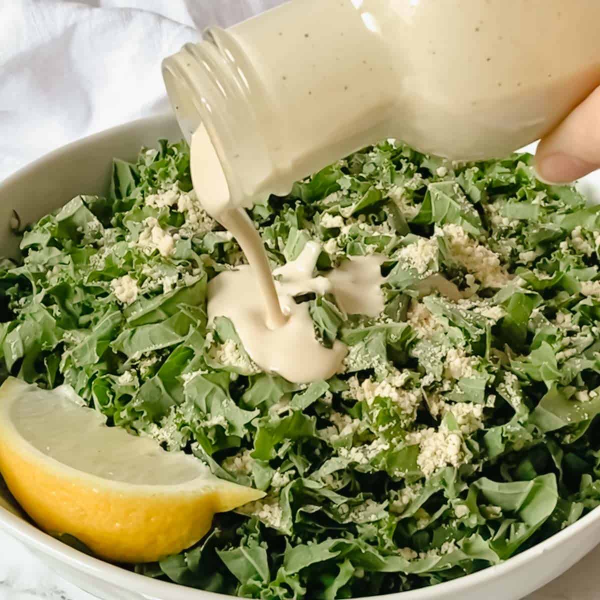 dressing being poured onto kale salad in white bowl.