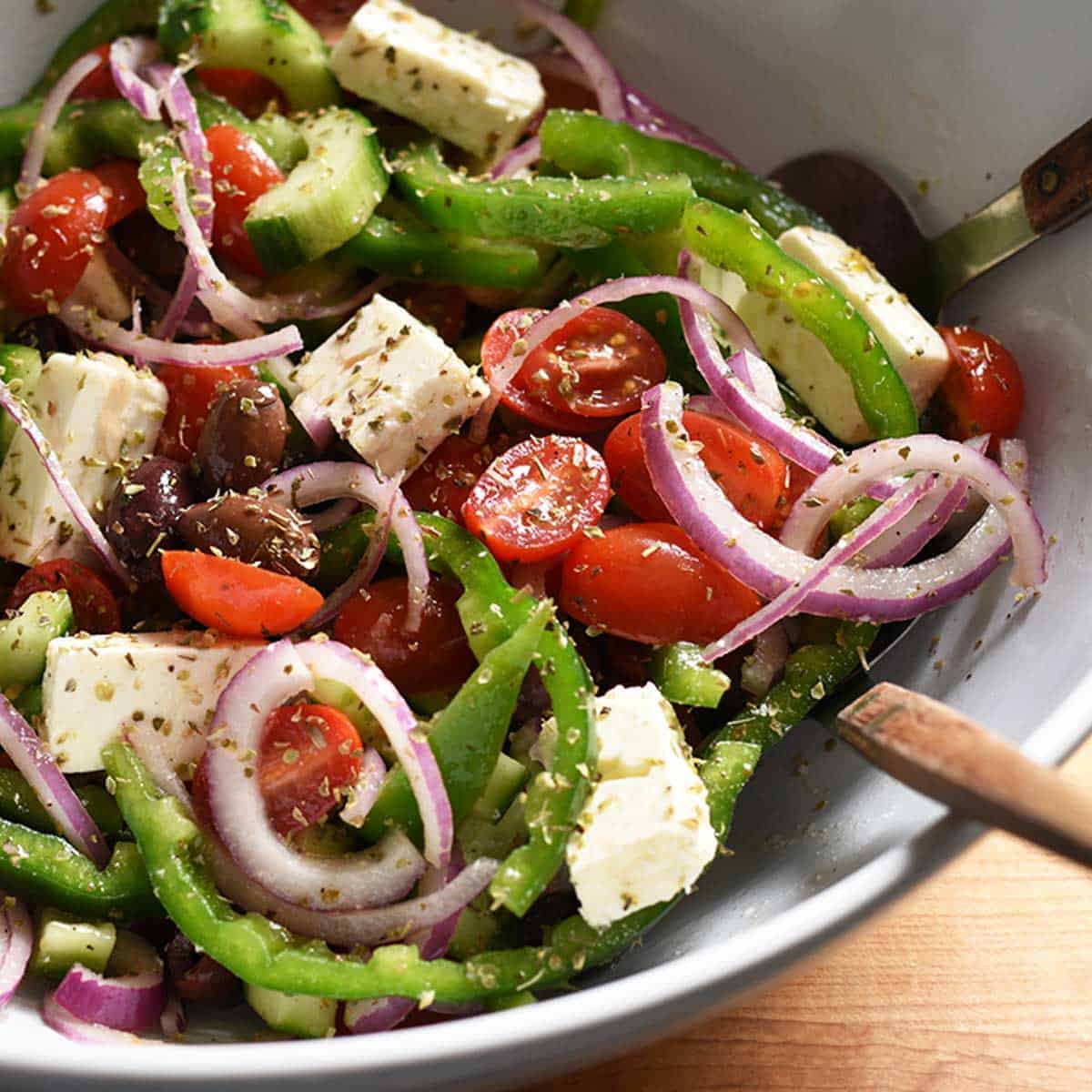 close up of greek salad in a bug white bowl with serving spoons.