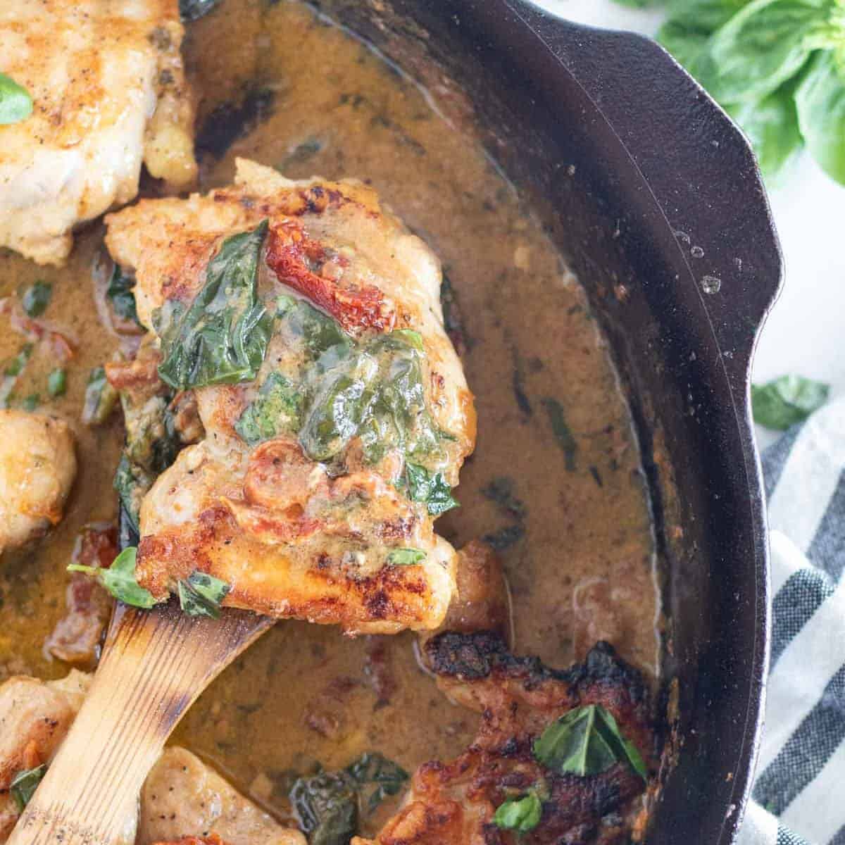 piece of tuscan chicken being held over skillet on a large wooden spoon.