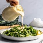 dairy free caesar dressing being poured over chopped greens on white plate.