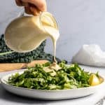 dairy free caesar dressing being poured over chopped greens on white plate.