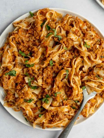 spaghetti bolognese on white plate sprinkled with basil with fork twirled in pasta taking a bite.